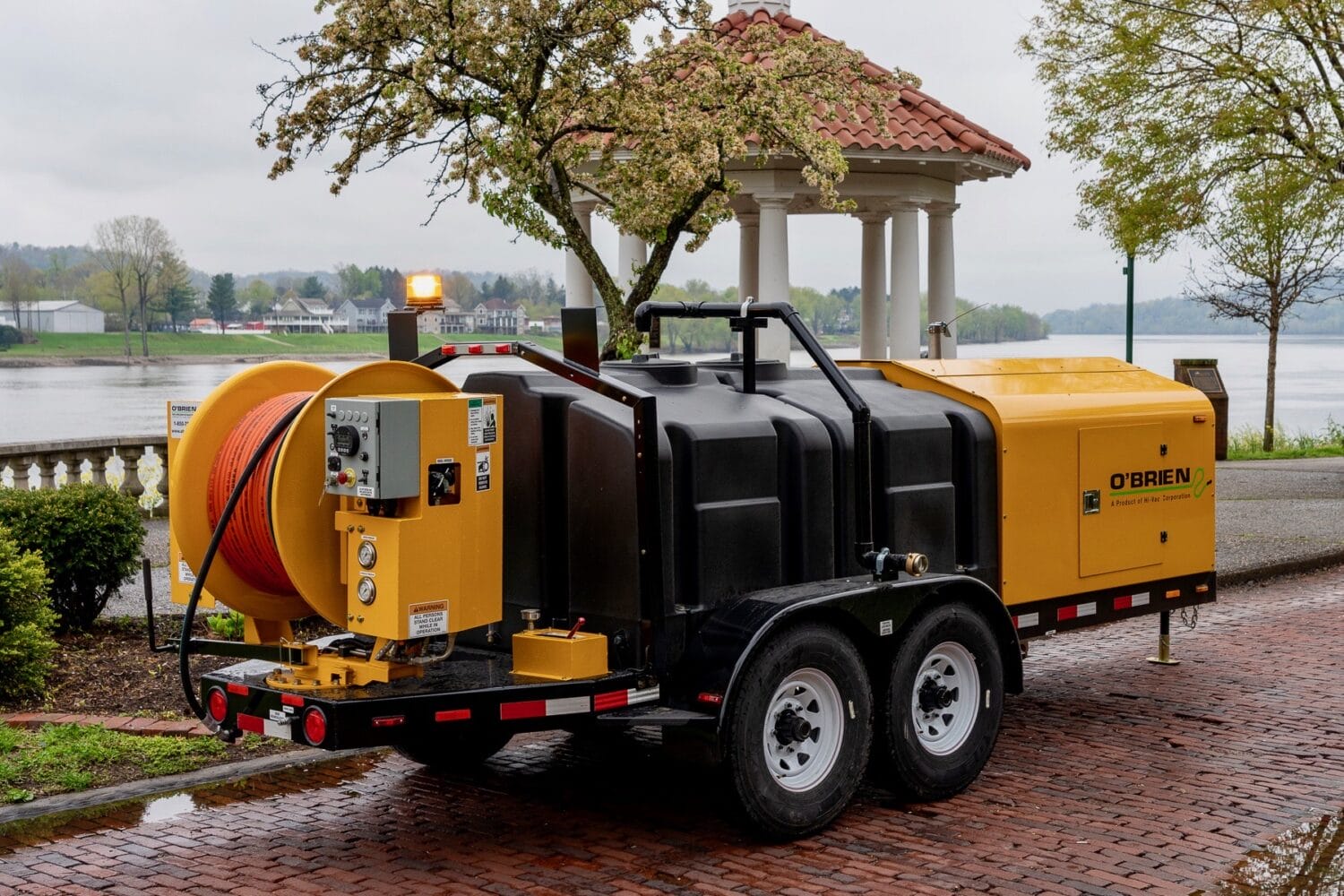 O'Brien Sewer Jetters trailer-mounted hydro jetter with orange hose reel and tandem axle parked by the river.
